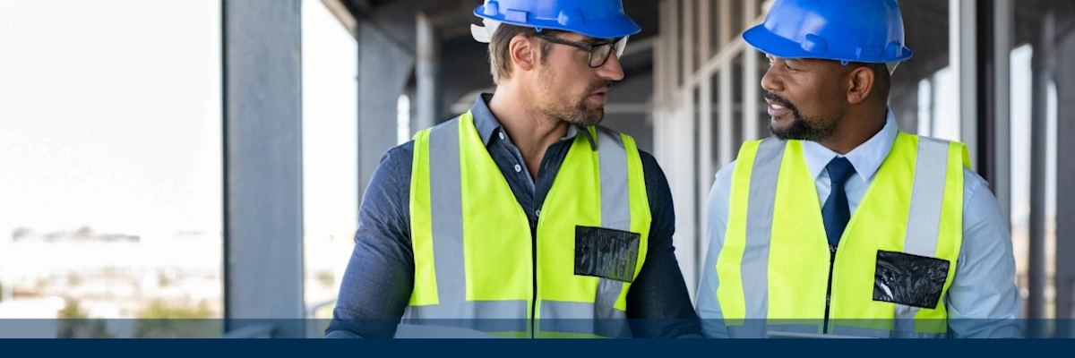 Two construction workers in safety vests and helmets discussing at a building site.