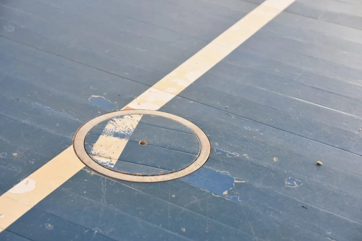 Close-up of a weathered blue wooden floor with a diagonal yellow stripe and an embedded circular metal plate.