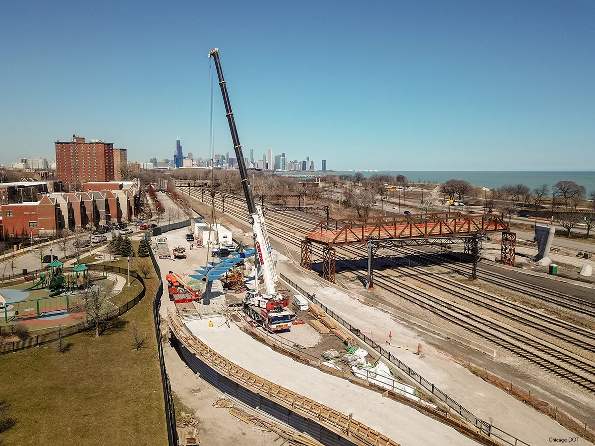A large crane is used to construct a pedestrian bridge over railroad tracks, with the Chicago skyline and Lake Michigan in the background.
