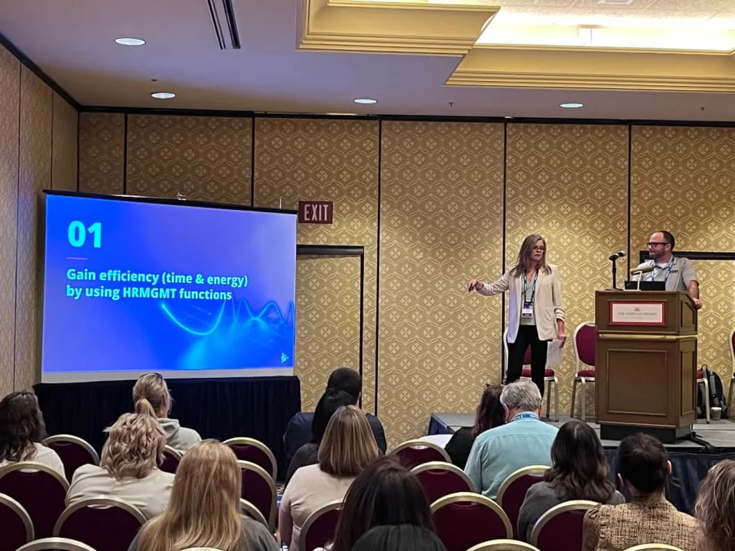 A woman stands on a stage with a large screen to the left of a podium, presenting to an audience about using HRMGMT functions
