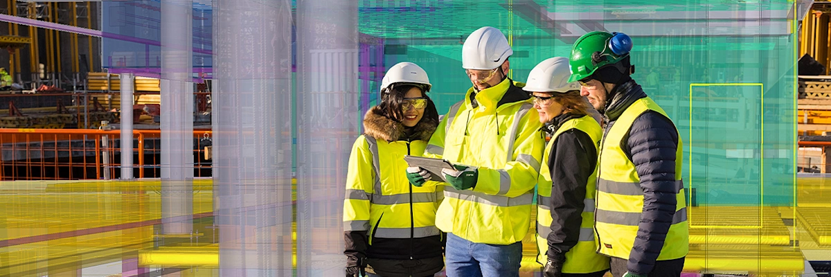 Four construction workers in safety gear review a digital tablet overlaying a colorful 3D building blueprint.