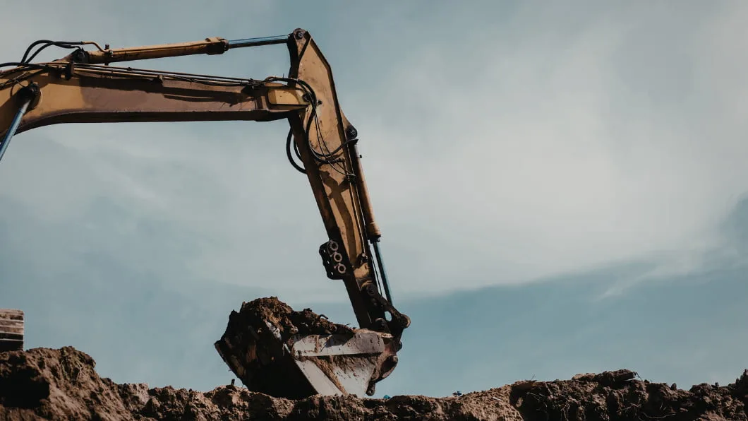 A yellow excavator arm lifting a bucket of dirt at a construction site against a blue sky.