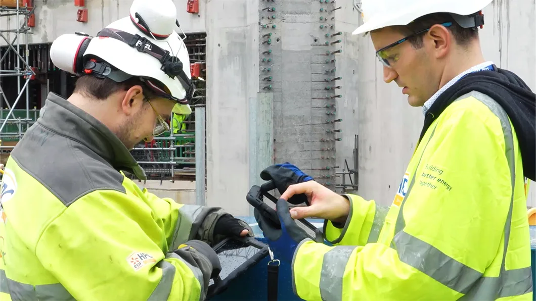 Two construction workers in yellow hi-vis jackets and white hard hats review a tablet device at a building site.