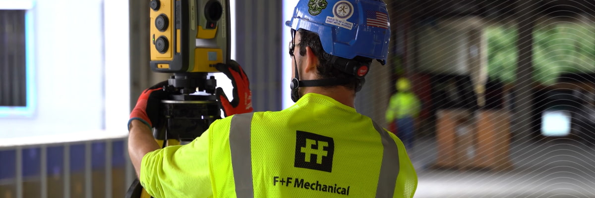 Worker in an F+F Mechanical safety vest operating a Trimble Robotic Total Station at a construction site.