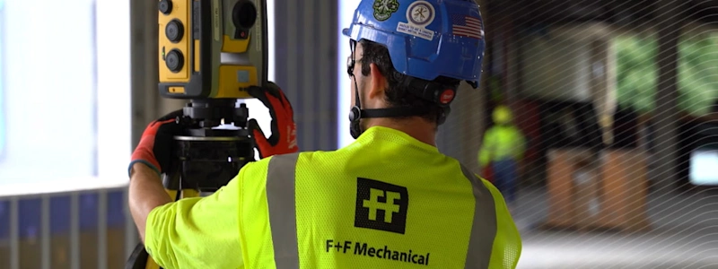 Worker in an F+F Mechanical safety vest operating a Trimble Robotic Total Station at a construction site.