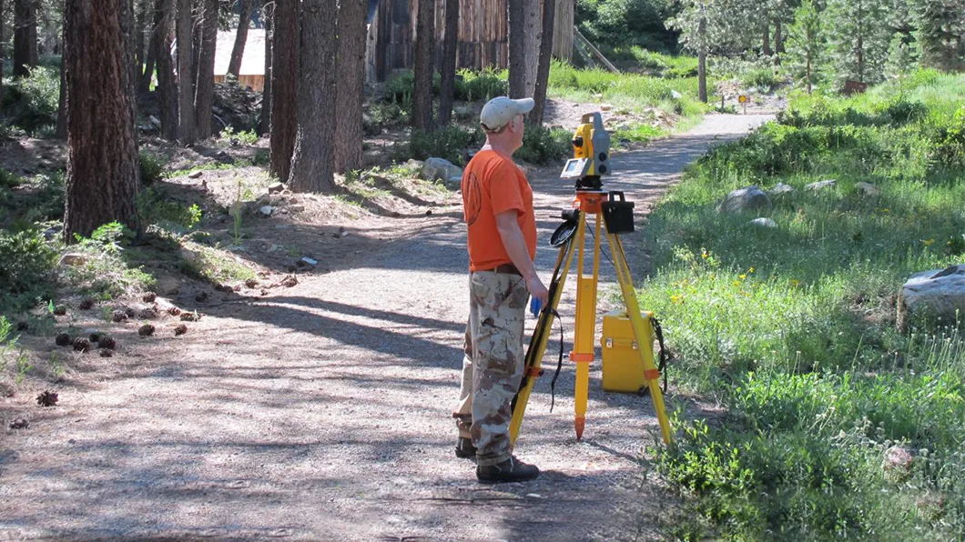 Person operating a yellow & grey total station on a tripod, with a yellow case, on a dirt path.