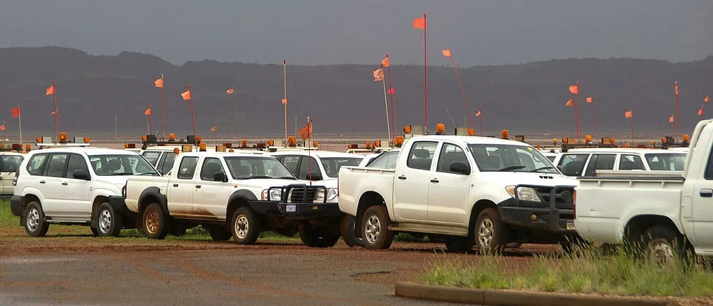 A fleet of white trucks and SUVs are parked in a field with orange flags on poles in the background.