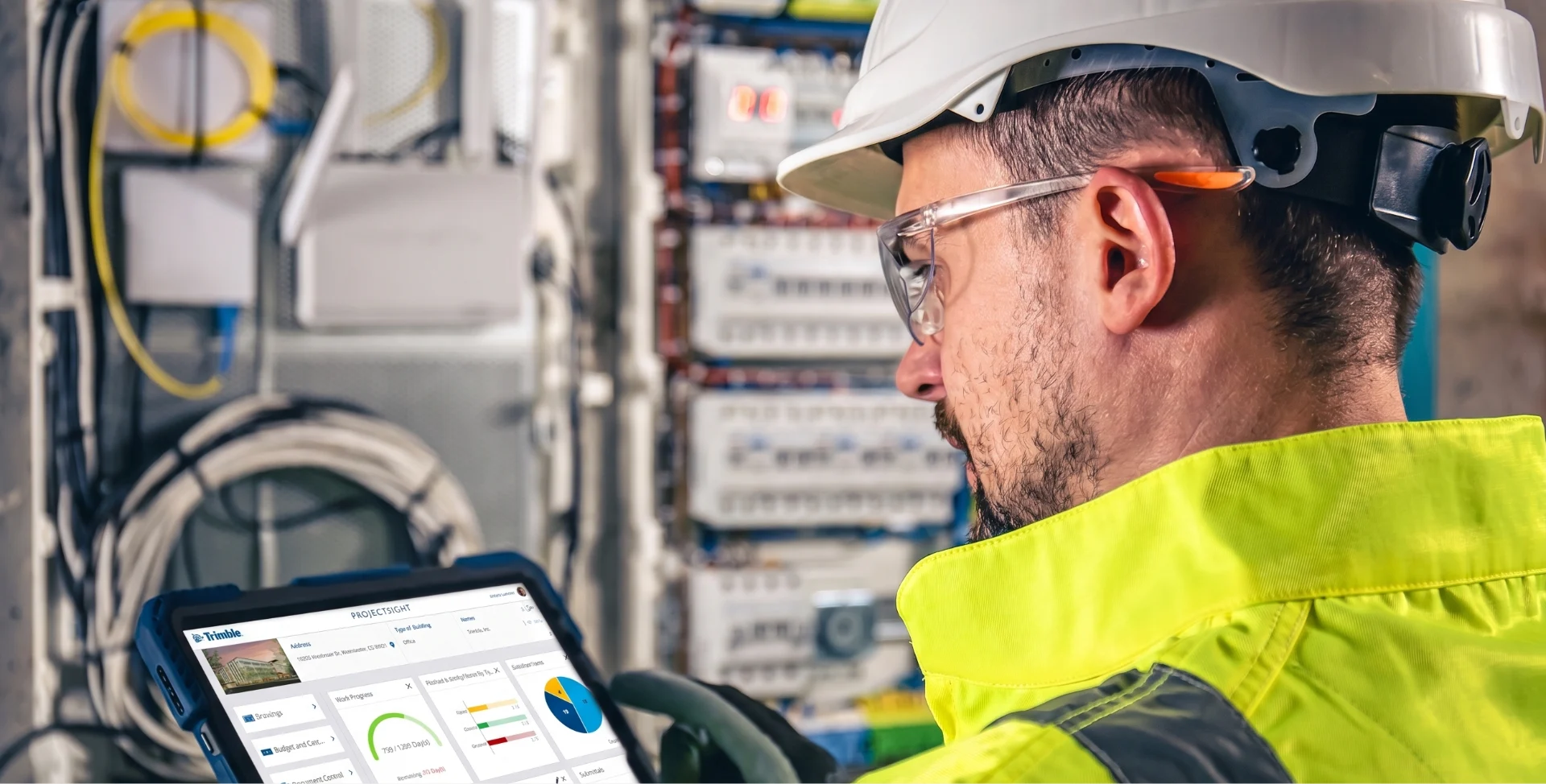 Electrician in a white hard hat and yellow vest reviews Trimble ProjectSight data on a tablet near an electrical panel.