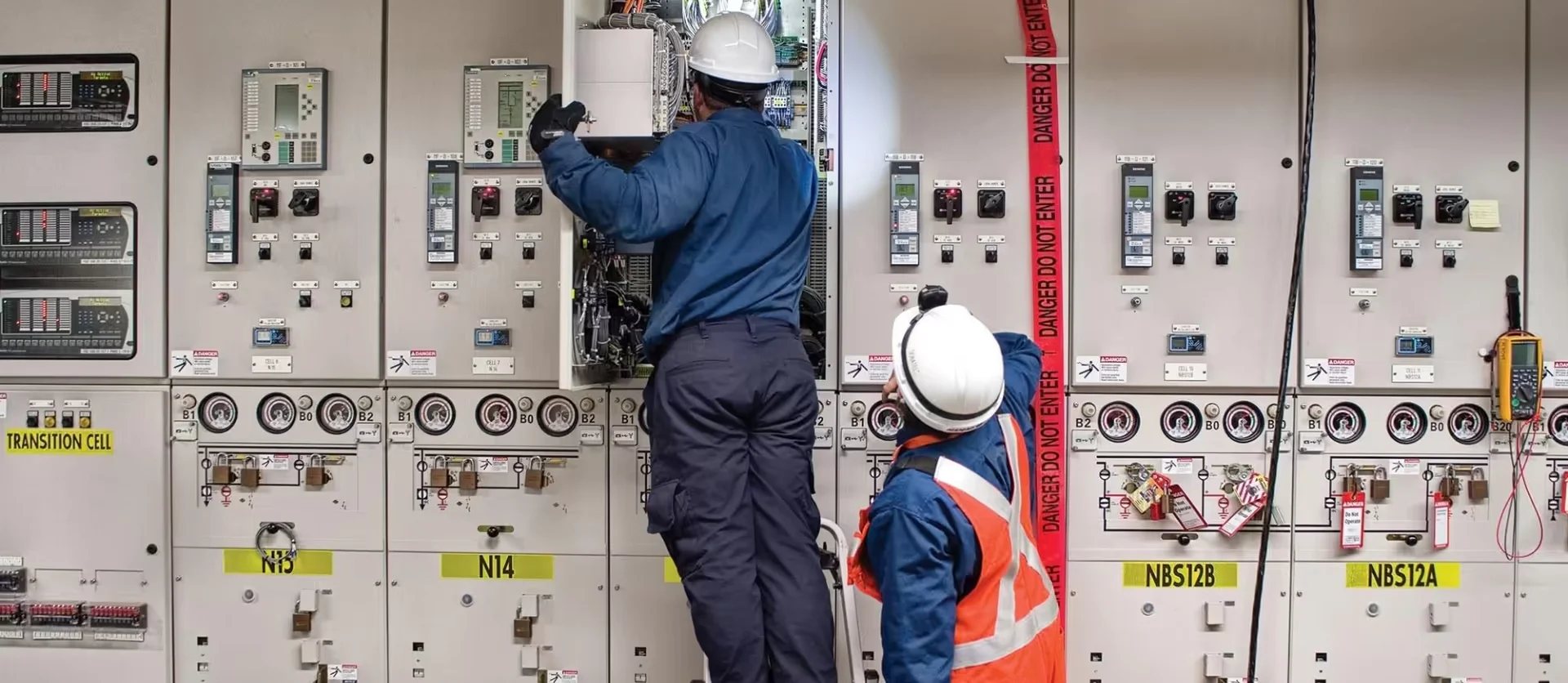 Two EPCOR electricians in hard hats inspecting wiring inside a large industrial electrical control panel.