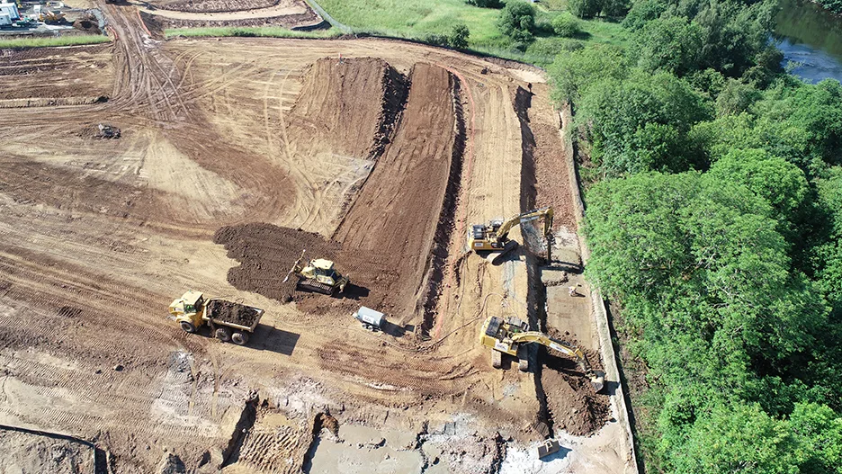 Construction site with excavators and dump trucks grading dirt next to a wooded area.