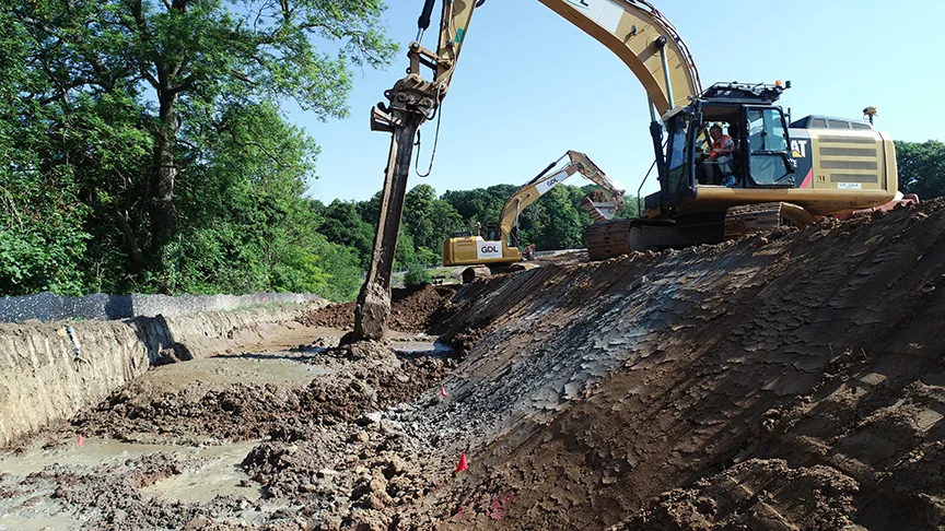 Excavators dig a trench on a sunny day with trees in the background.