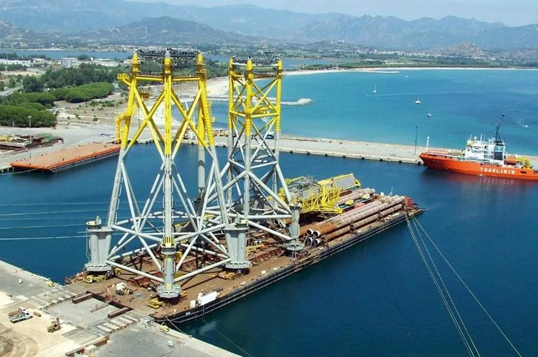 A large offshore wind turbine jacket is being transported on a barge, with an orange tugboat nearby.