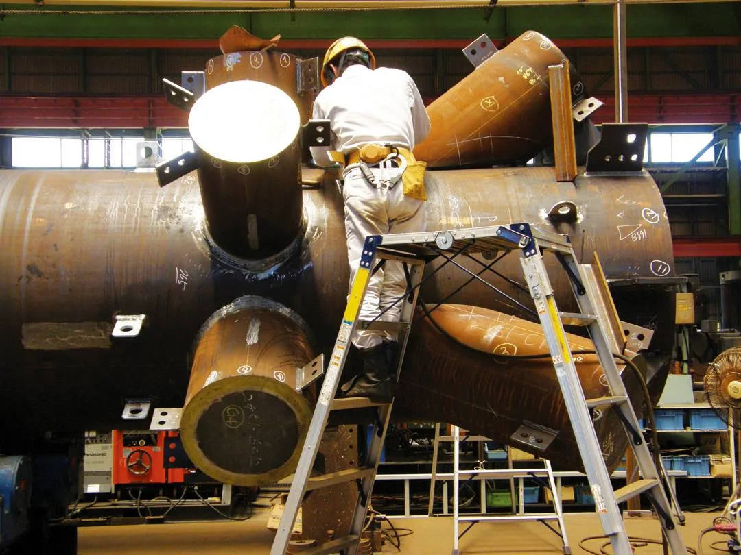A worker in a hard hat and white overalls stands on a ladder assembling a large, complex metal structure in a factory.
