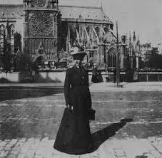 Woman in a long dress and hat stands in front of Notre Dame Cathedral.