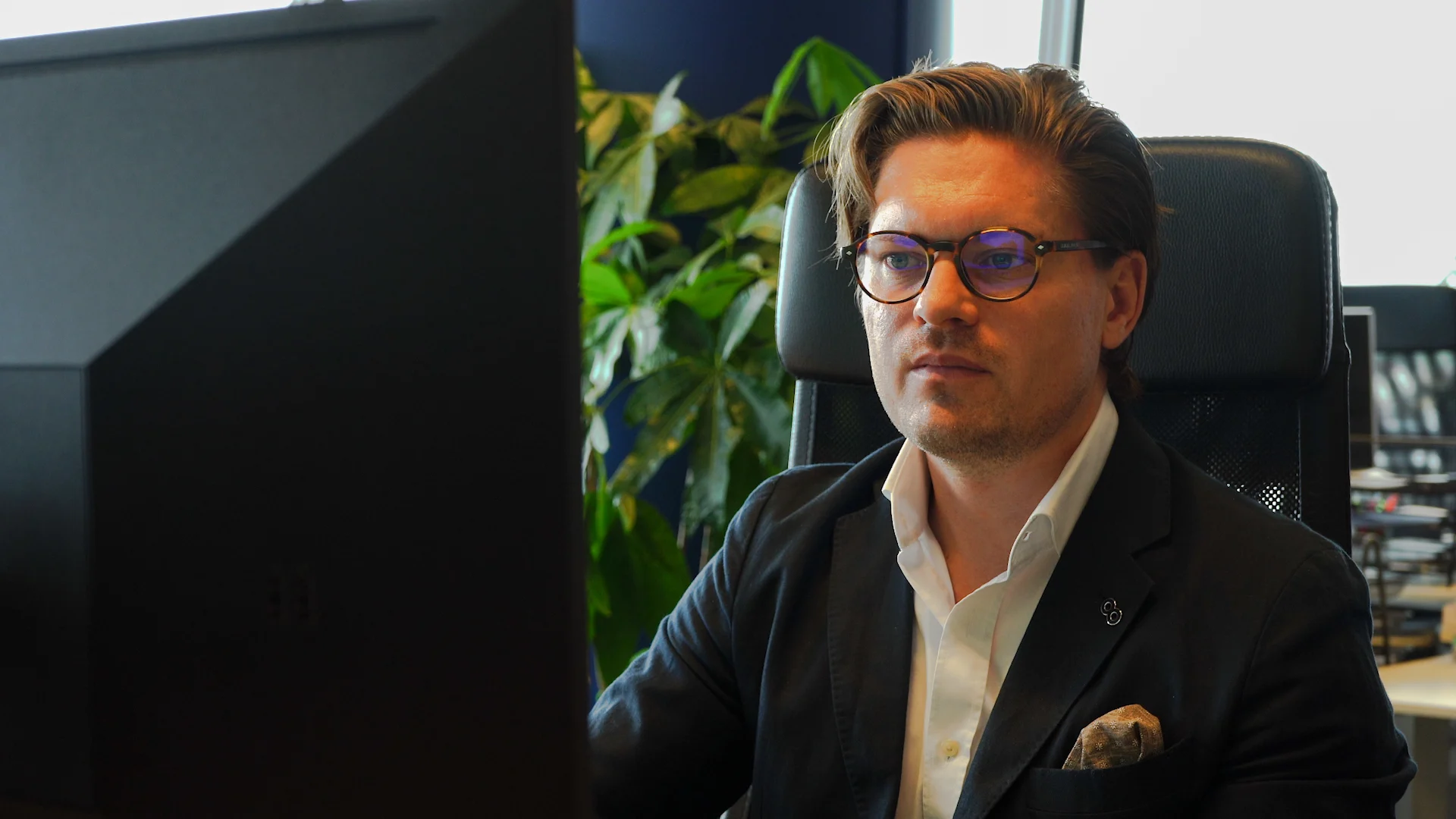 A man in a dark suit and glasses looks intently at a computer screen, seated at an office desk.