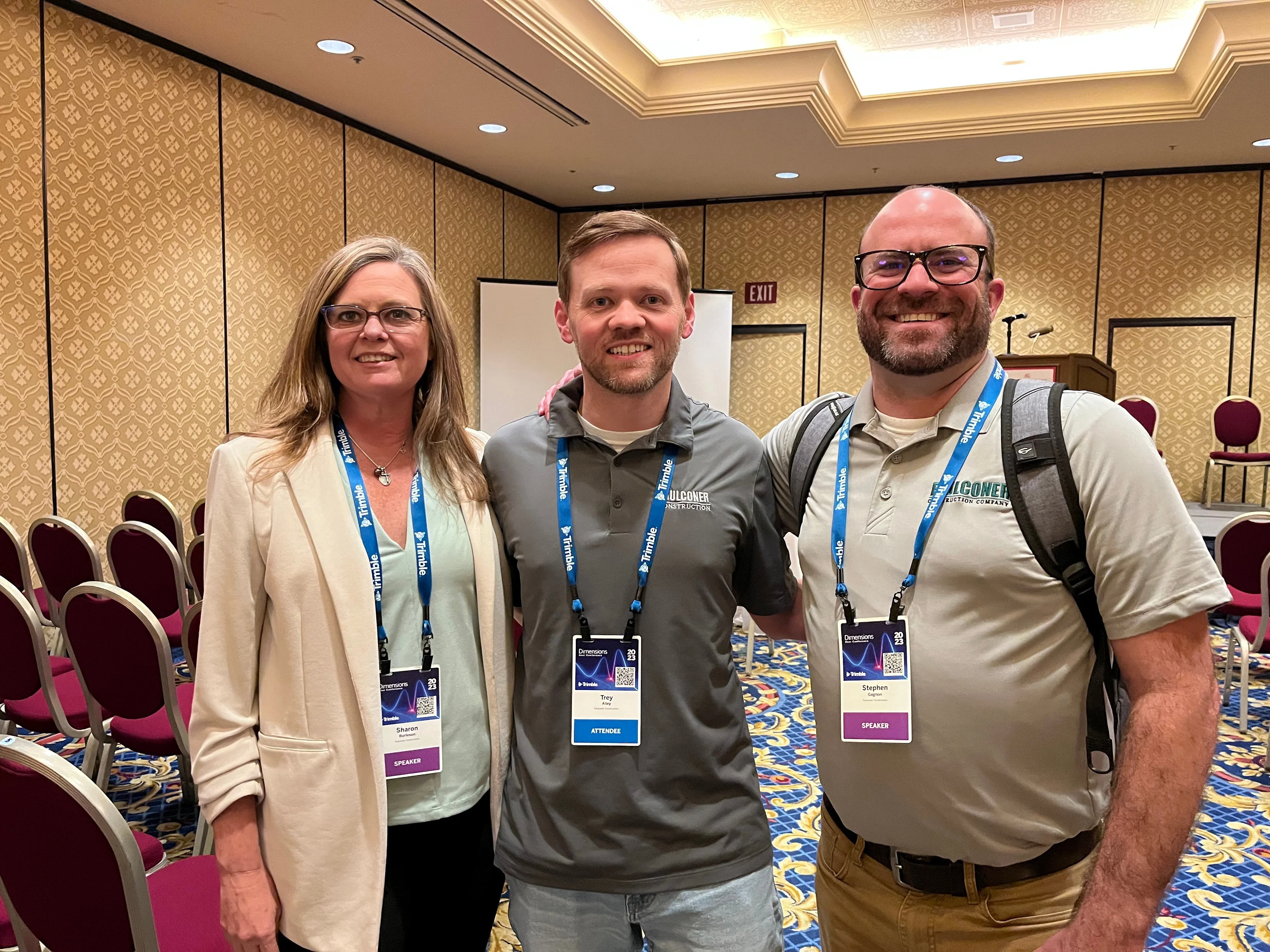 Three individuals, pose for a photo in front of a conference room backdrop, all wearing lanyards with name tags.