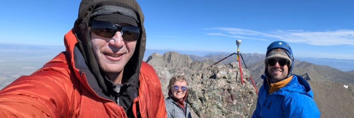 Three hikers smiling at a mountain summit selfie, wearing winter gear, with rocky peaks and a clear blue sky behind them.
