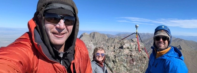 Three hikers smiling at a mountain summit selfie, wearing winter gear, with rocky peaks and a clear blue sky behind them.