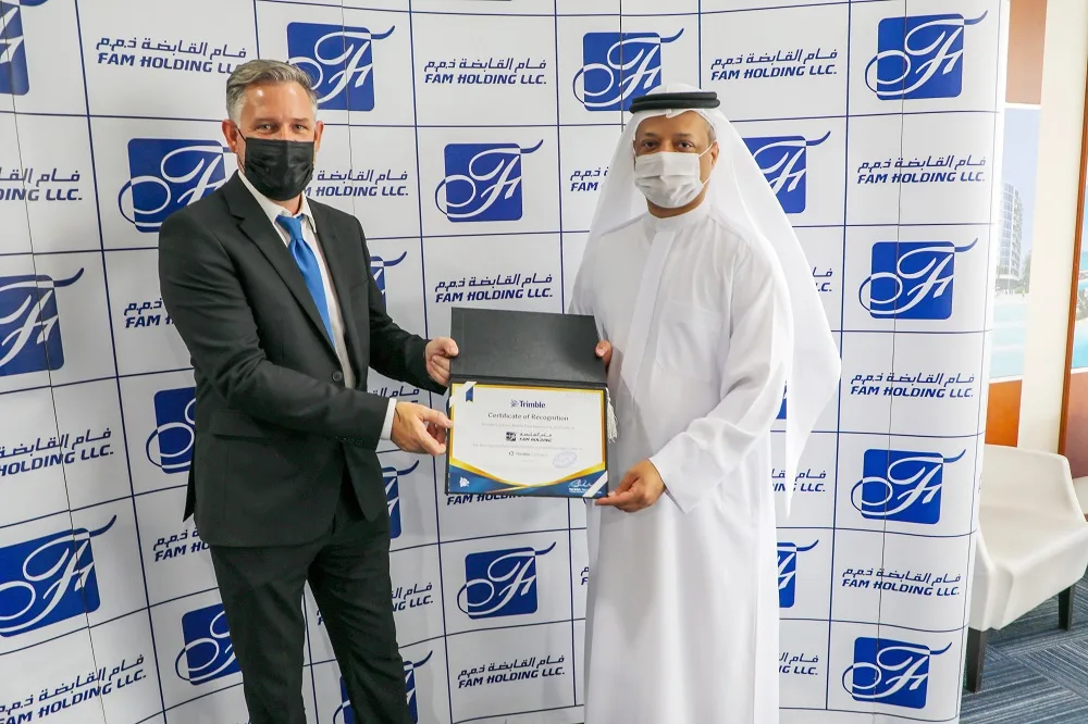 Two men, one in a suit and one in a white thobe, exchange a framed "Trimble Certificate of Recognition."