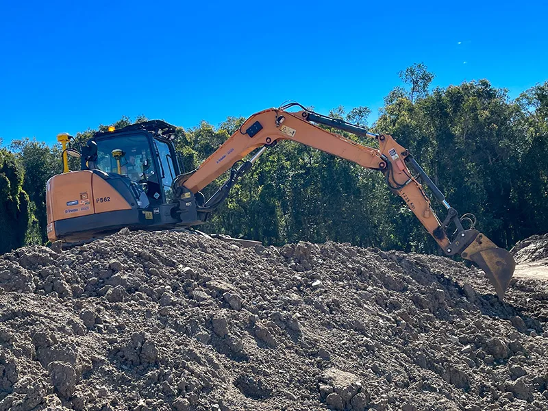 Orange excavator on a pile of dirt against a blue sky and trees.
