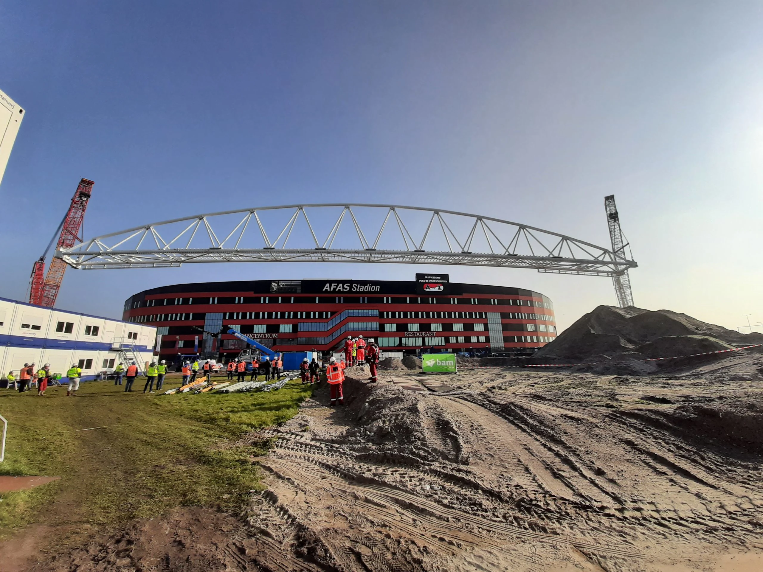 Construction workers in hard hats gather near a large stadium under construction, with a massive steel truss being lifted into place by cranes.