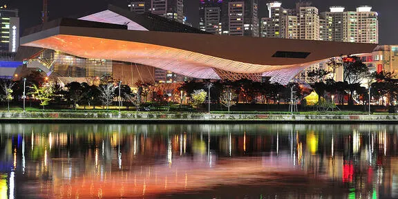 The Busan Cinema Center, illuminated at night, with its distinctive, sweeping roof structure, reflected in the calm waters of the canal, in the city.