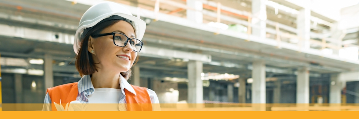 A person at a construction site wearing a hard hat and safety vest, holding a tablet and blueprints.