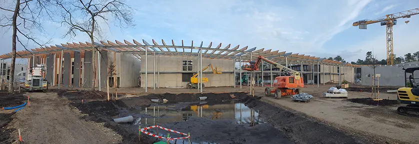 Construction of a long, modern building with a wooden-beam roof structure, concrete walls, and glass windows, surrounded by a muddy construction site.