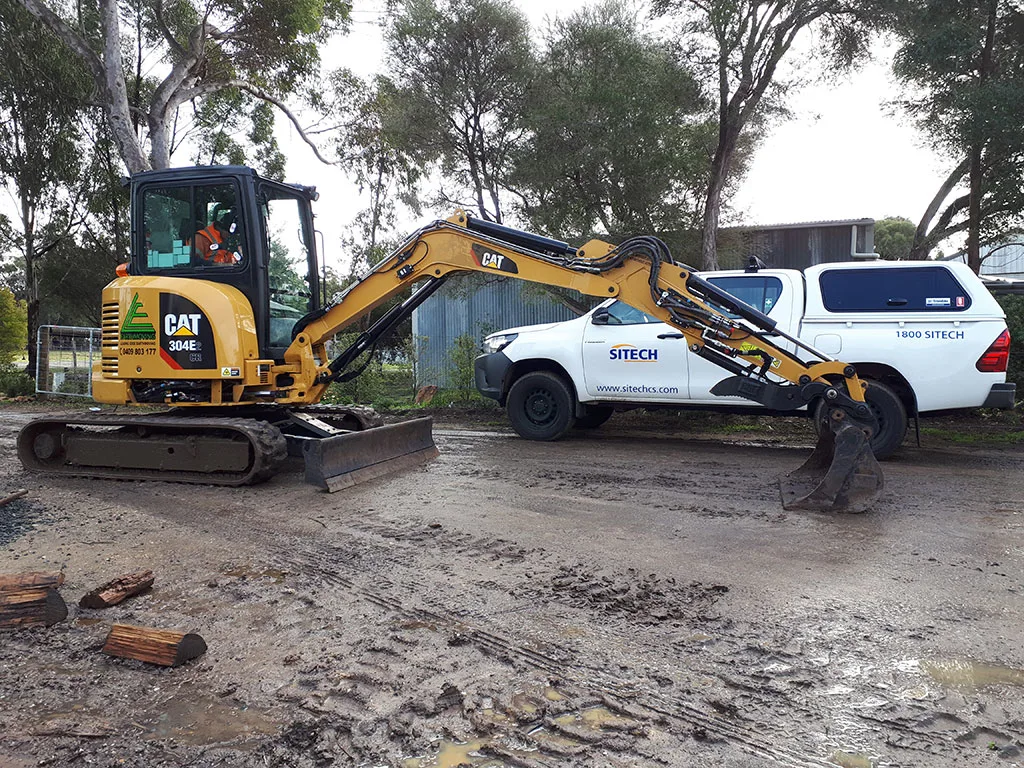 A yellow CAT 304E mini excavator is parked on muddy ground next to a white Sitech truck.