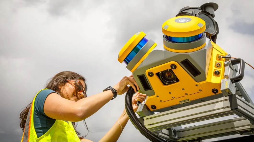 A woman in a yellow safety vest adjusts a yellow and gray surveying or scanning device mounted on a rack outdoors.