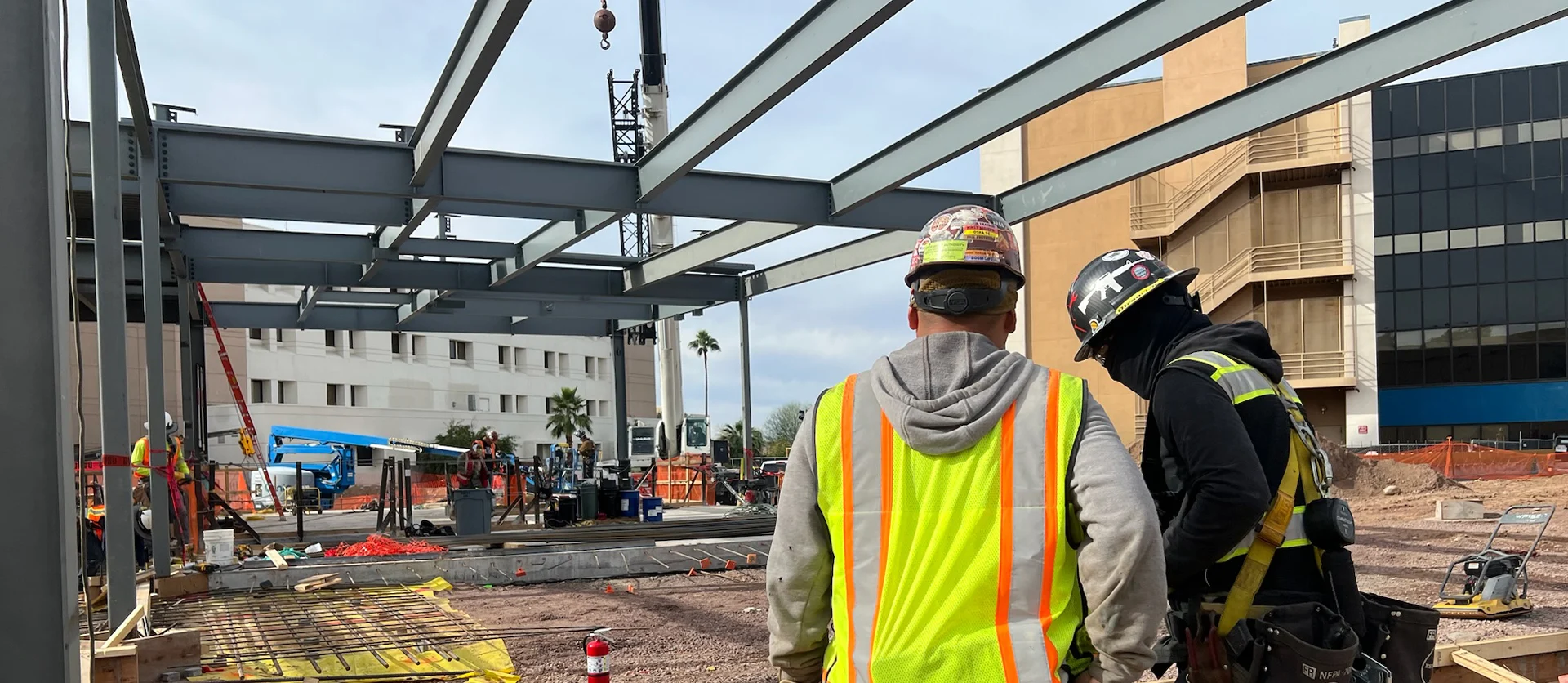 Two construction workers in hard hats and safety vests observe the progress of a large steel building framework on a busy job site.