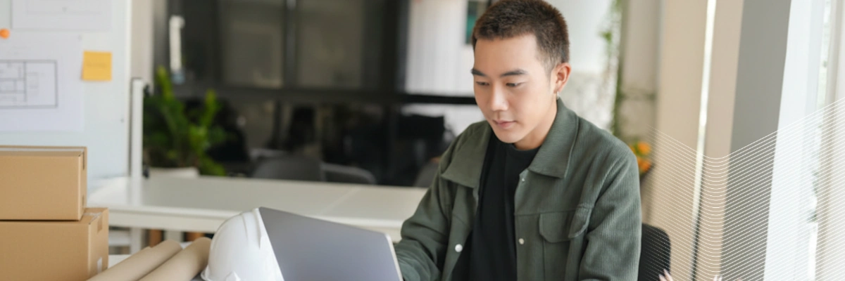 Person in green jacket working on laptop in bright modern office space with plants visible in background.