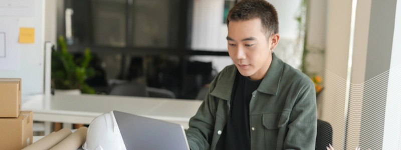 Person in green jacket working on laptop in bright modern office space with plants visible in background.