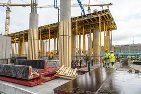 Construction workers in hard hats and safety vests stand around a concrete foundation with large cylindrical columns and yellow scaffolding.