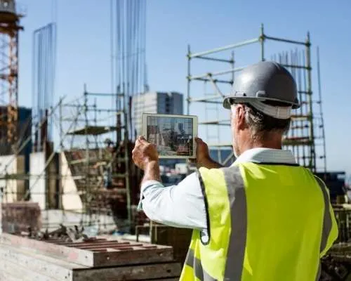 Construction worker in safety gear using tablet to monitor building progress at urban construction site.