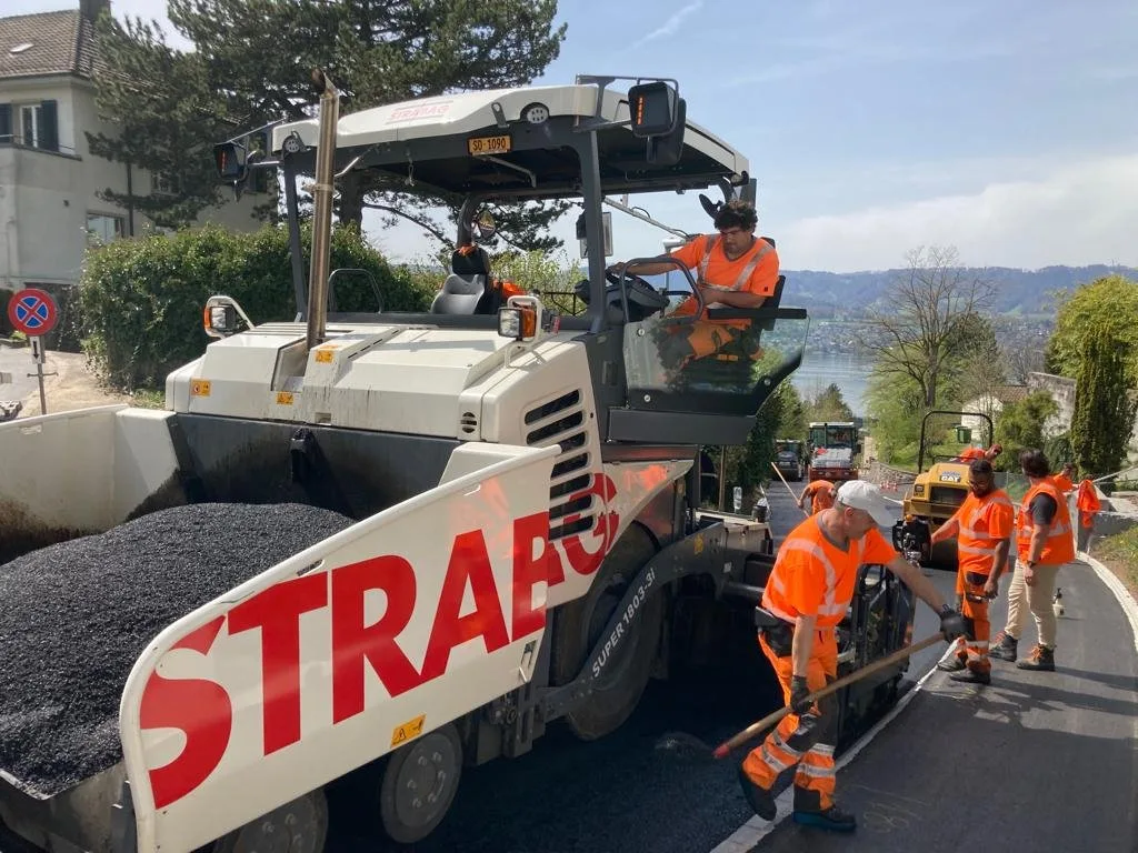 Road construction crew in orange safety gear operates heavy machinery, laying asphalt on a sunny day near a residential area