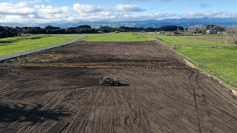 Construction grader levels a large, cleared dirt field with green fields and mountains in the background.
