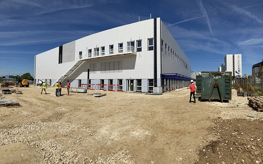 Construction site with a large white building under development, workers in hard hats, and heavy machinery.