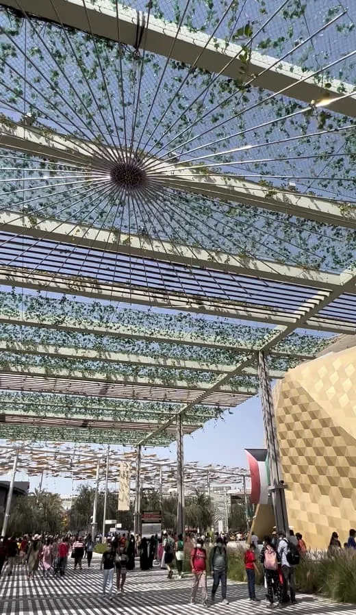 A canopy of metal and vines covers a plaza with people walking beneath.