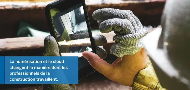 A construction worker in gloves holds a smartphone, showing a digital representation of a construction project.