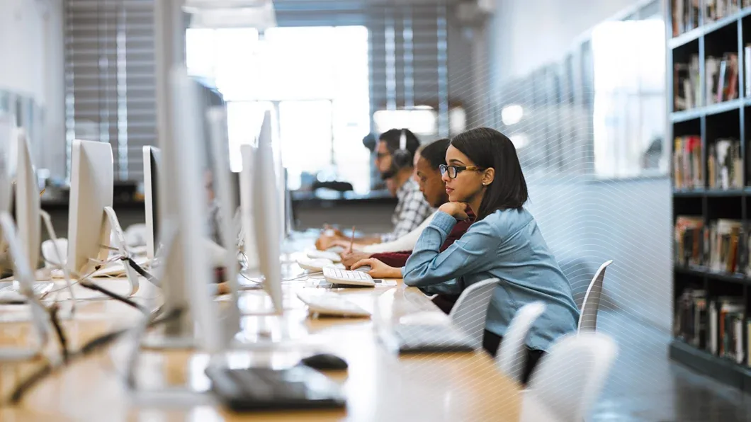 A woman with glasses and several other individuals work at a row of computer stations in a bright, modern library or computer lab.