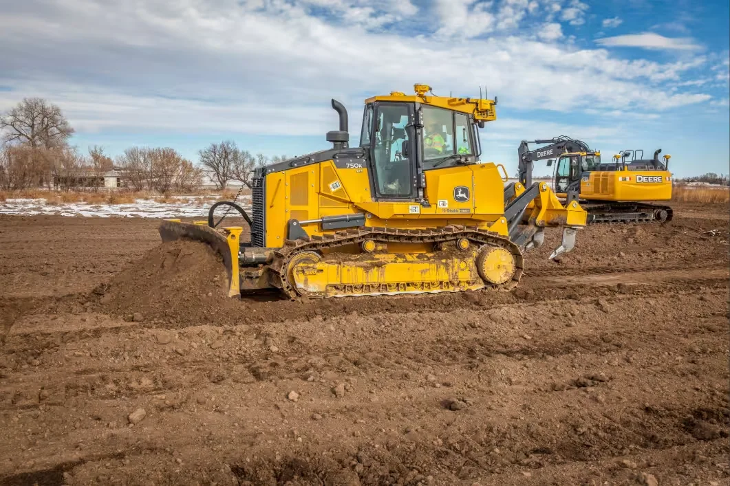 A yellow John Deere bulldozer pushes dirt on a construction site, with an operator in the cab and an excavator in the background.