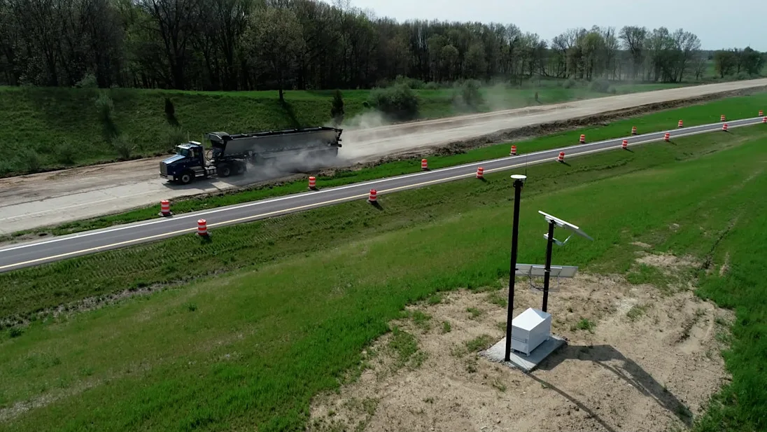 A truck dumps asphalt on a new road construction site, with traffic cones and survey equipment visible.