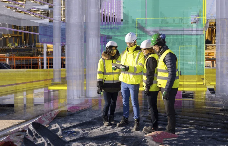 Four construction workers in hard hats and safety vests look at a tablet displaying a digital model of a building overlayed on a construction site.