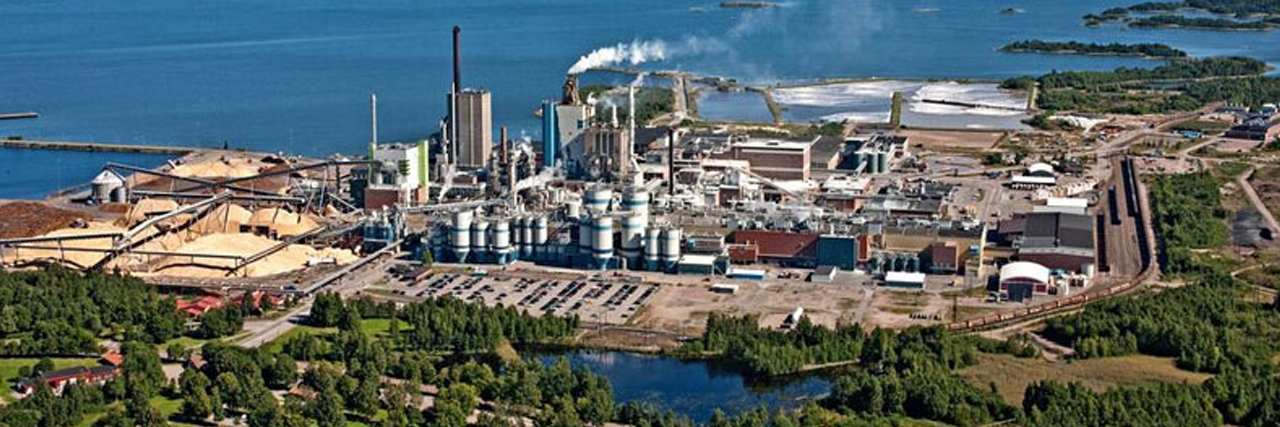Aerial view of an industrial plant by a large body of water, emitting white smoke. Greenery contrasts with the structures.