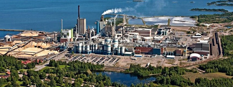 Aerial view of an industrial plant by a large body of water, emitting white smoke. Greenery contrasts with the structures.