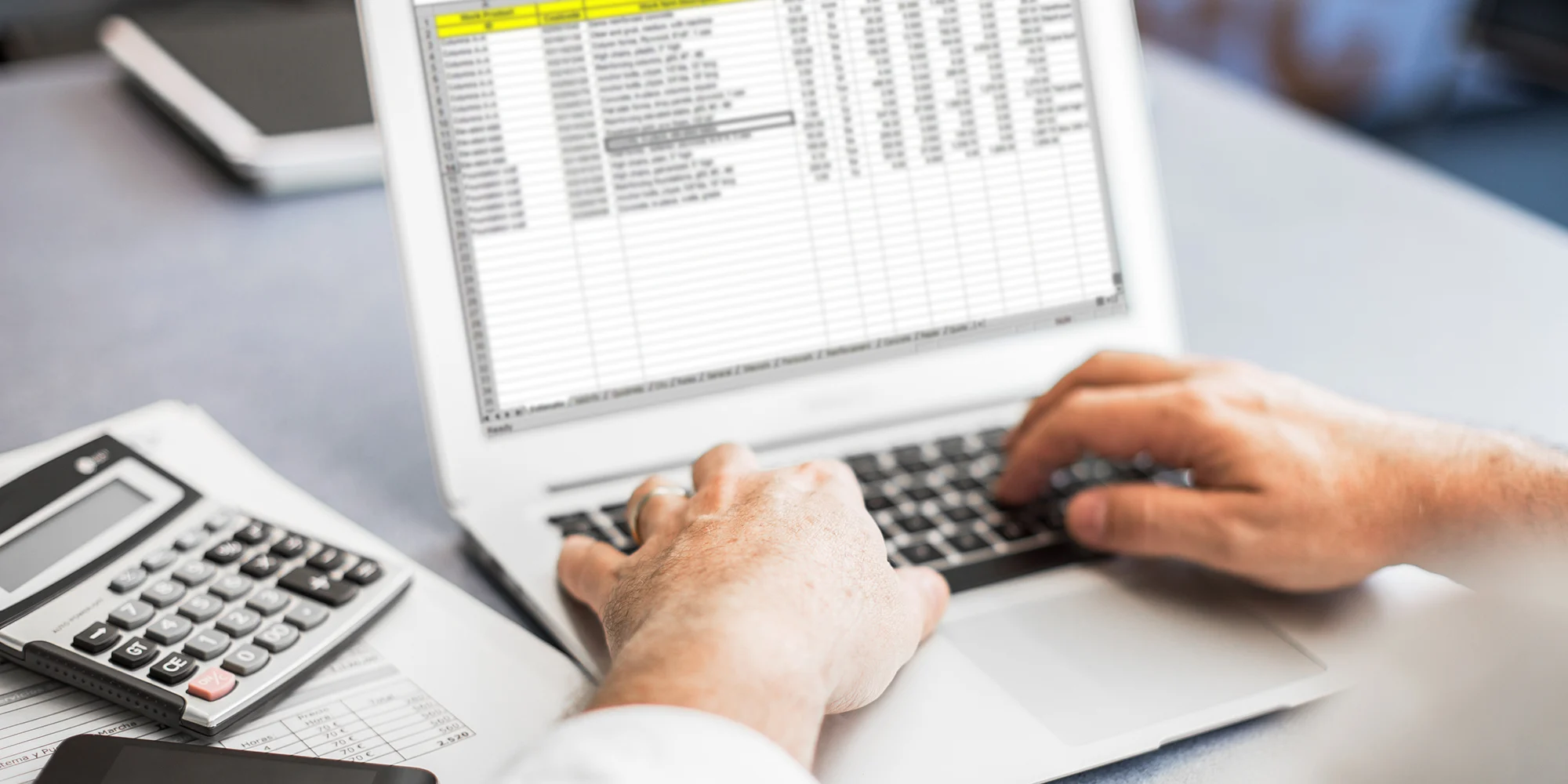 A person is using a laptop with a spreadsheet open, with a calculator and phone on the desk.