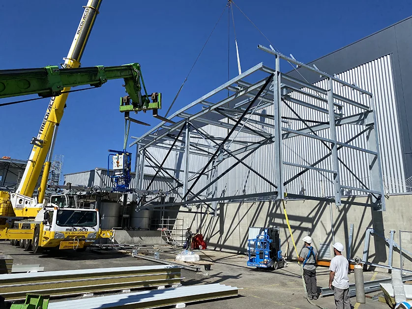 Workers use a crane and a lift to install a steel structure next to a modern building.