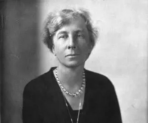 Portrait of a woman with short, wavy hair, wearing a pearl necklace and dark clothing.