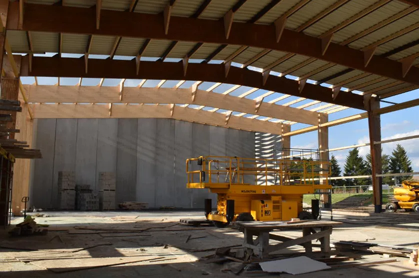 Construction of a large wooden-beamed building with concrete walls, showing a yellow scissor lift in the foreground.